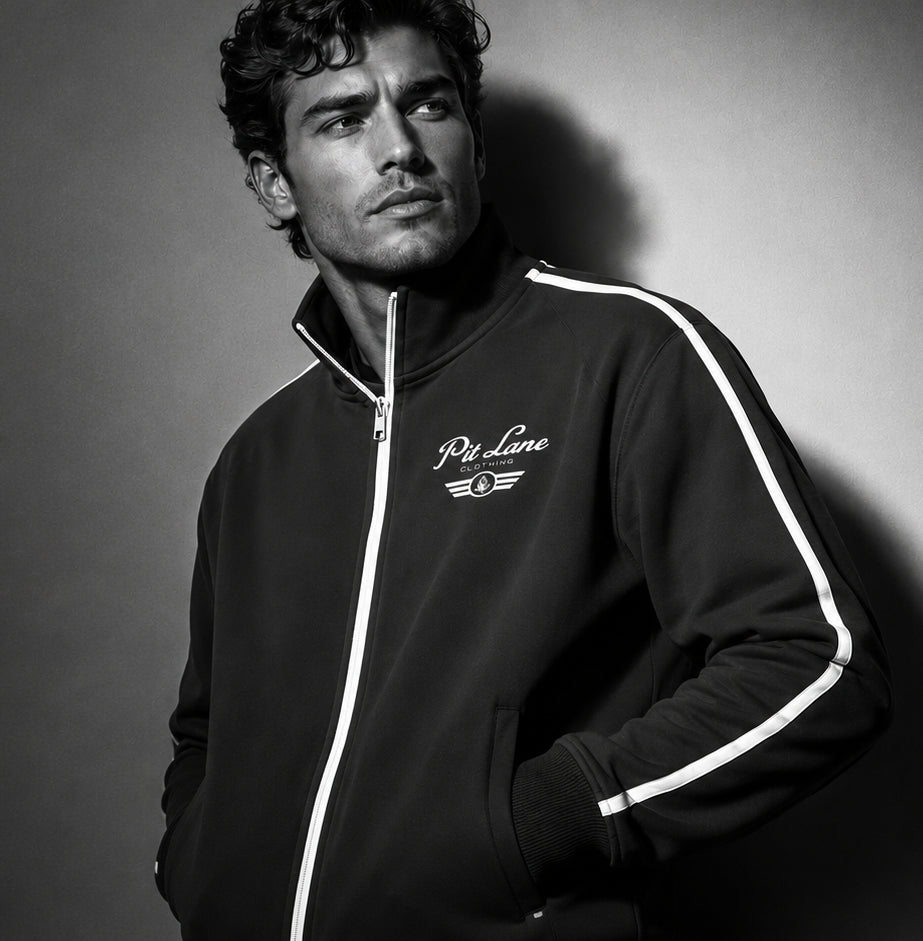 A man wearing a black zip-up track jacket with white stripe details and a white 'PIt Line Clothing' logo on the chest, photographed in a dramatic black-and-white studio setting.