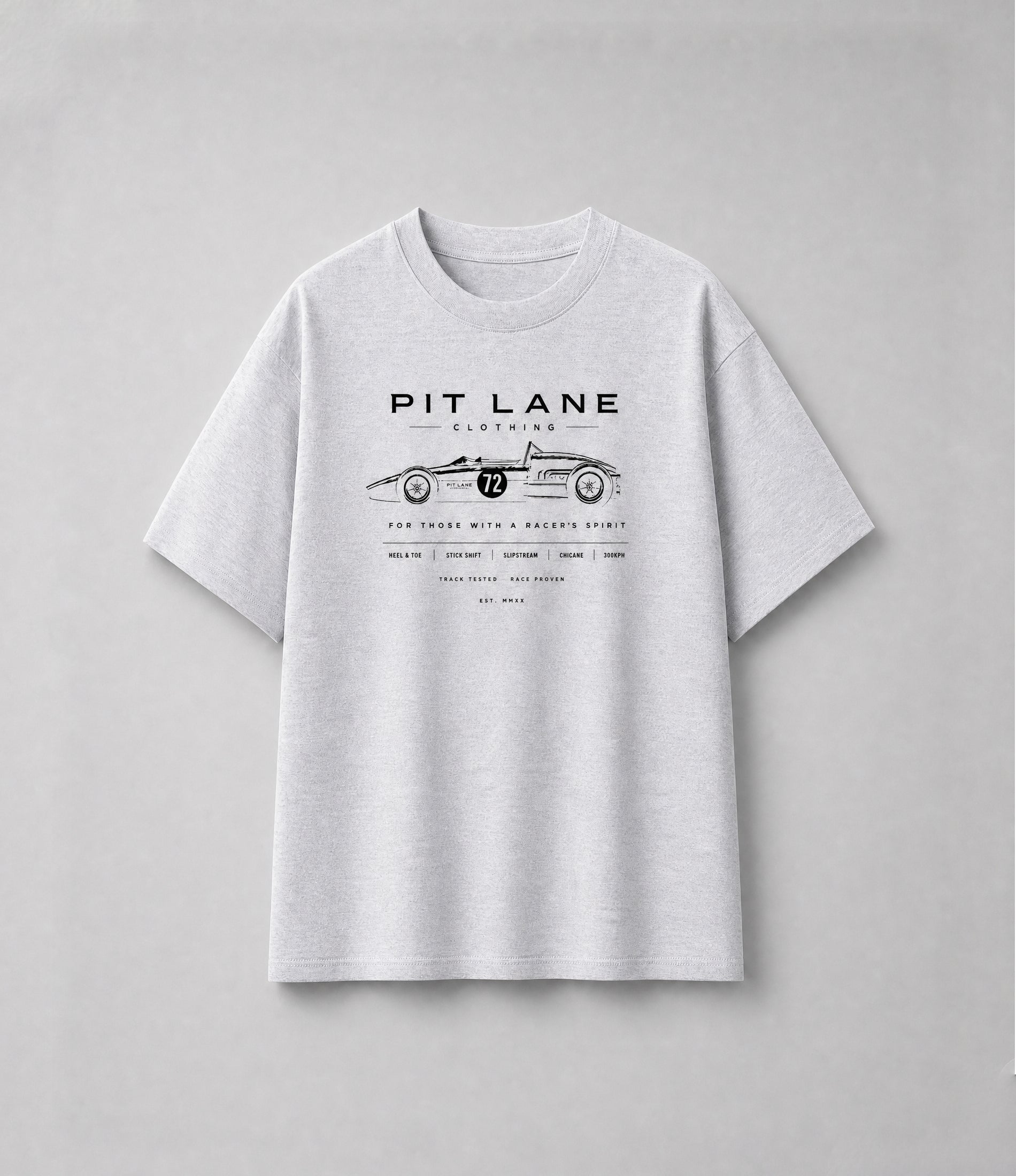 A man wearing a white short-sleeve graphic t-shirt with a brown geometric design and text reading 'PIT LANE CLOTHING' and 'APEX', paired with dark jeans, standing against a textured gray studio background.