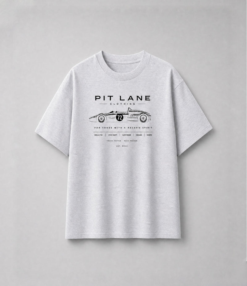 A man wearing a white short-sleeve graphic t-shirt with a brown geometric design and text reading 'PIT LANE CLOTHING' and 'APEX', paired with dark jeans, standing against a textured gray studio background.
