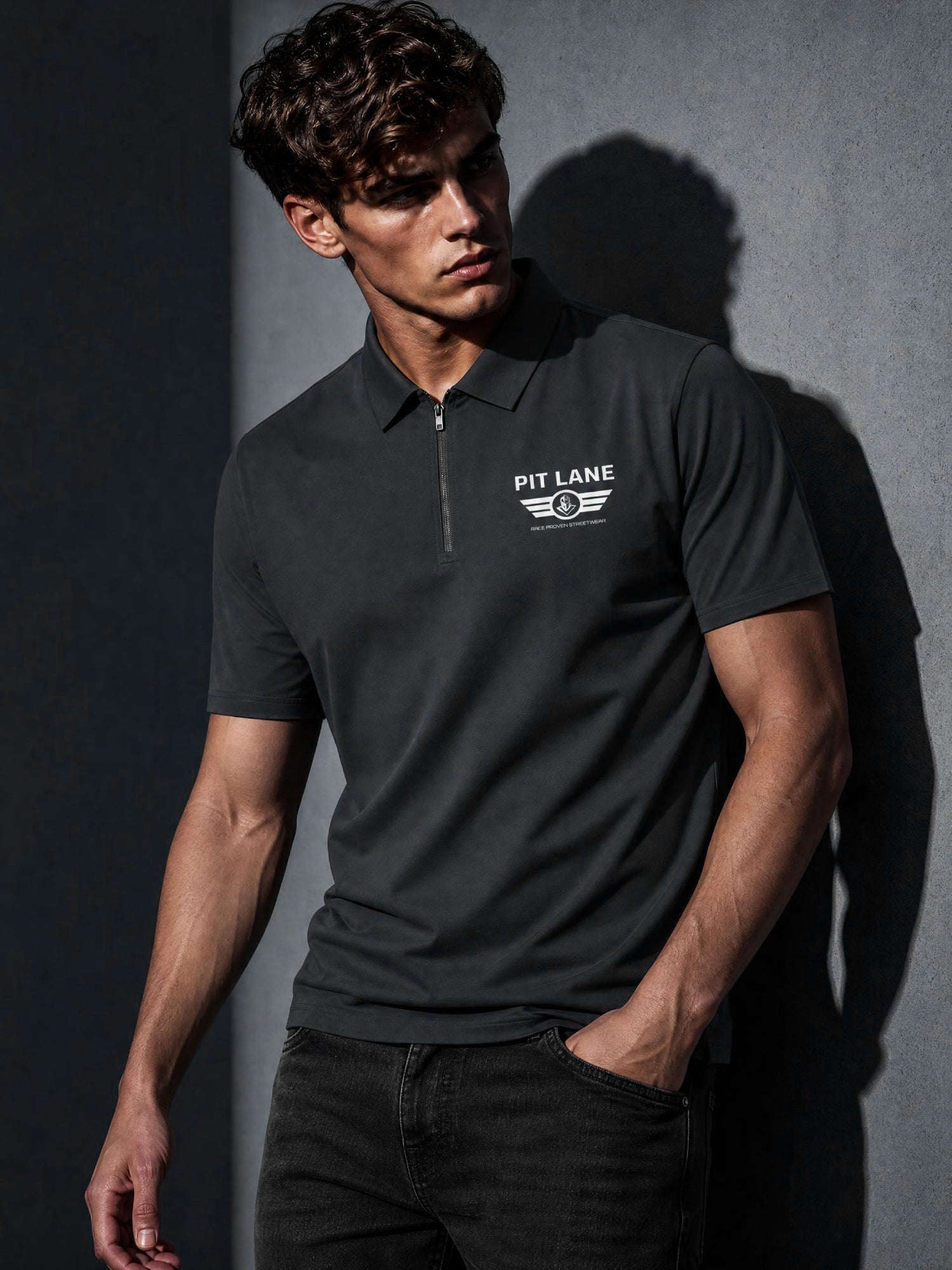 Man wearing grey Pit Lane Clothing polo shirt leaning against a grey wall