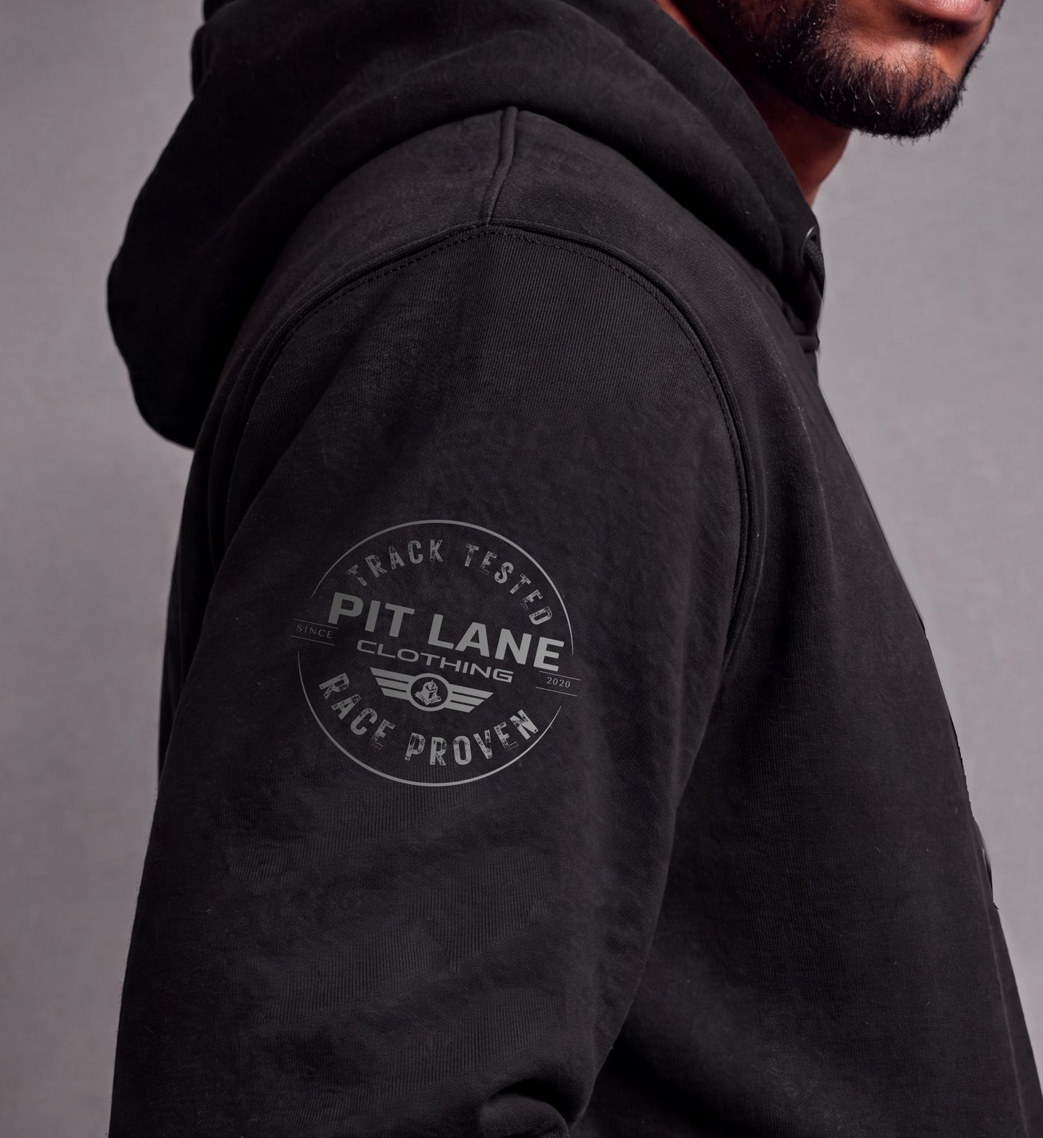 Close-up of a man wearing a black hooded sweatshirt, showing the side profile and a circular gray graphic on the upper left sleeve that reads 'PIT LANE CLOTHING' with 'TRACK TESTED' and 'RACE PROVEN'.