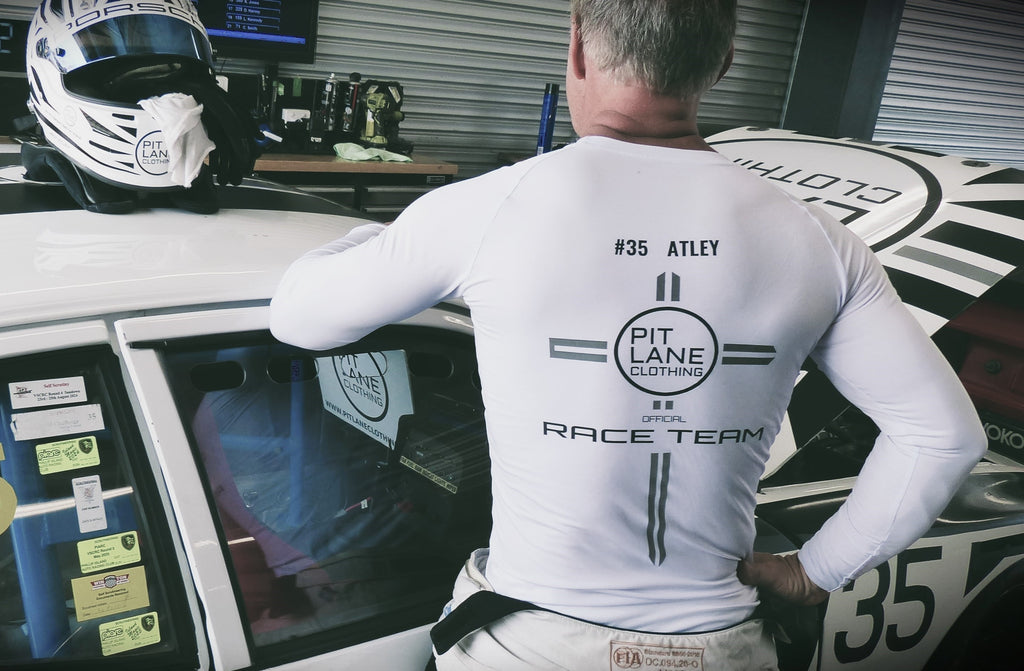 A man wearing a white long-sleeve race team shirt with 'PIT LANE CLOTHING' and '#35 ATLEY' printed on the back, leaning against a white race car in a garage setting.