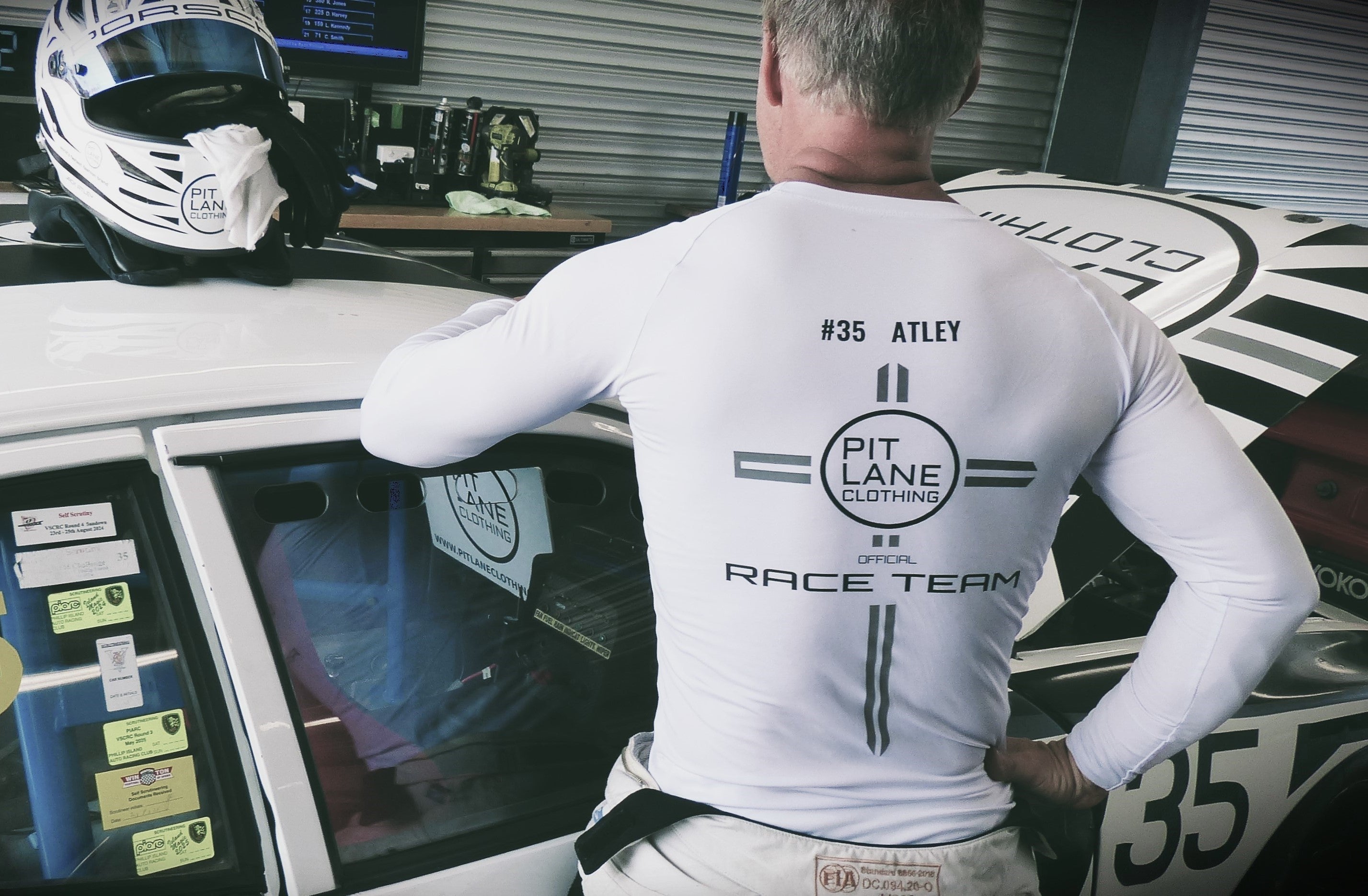 A man wearing a white long-sleeve race team shirt with 'PIT LANE CLOTHING' and '#35 ATLEY' printed on the back, leaning against a white race car in a garage setting.