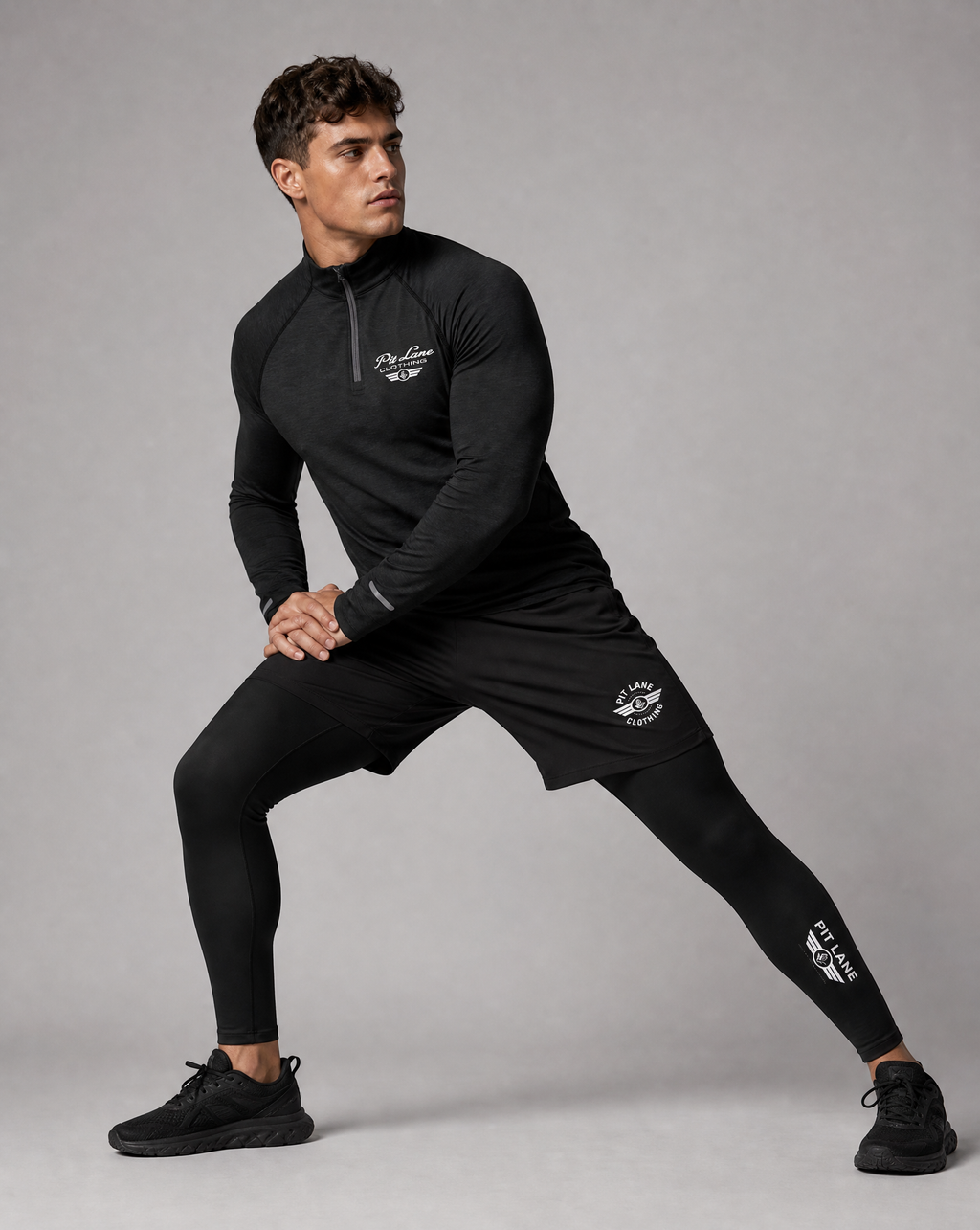 A man in a black PIT LANE CLOTHING athletic half-zip pullover, matching shorts, and full-length leggings poses in a side lunge against a solid gray studio background. The brand name 'PIT LANE CLOTHING' is printed in white on the chest, shorts, and lower leg of each garment.