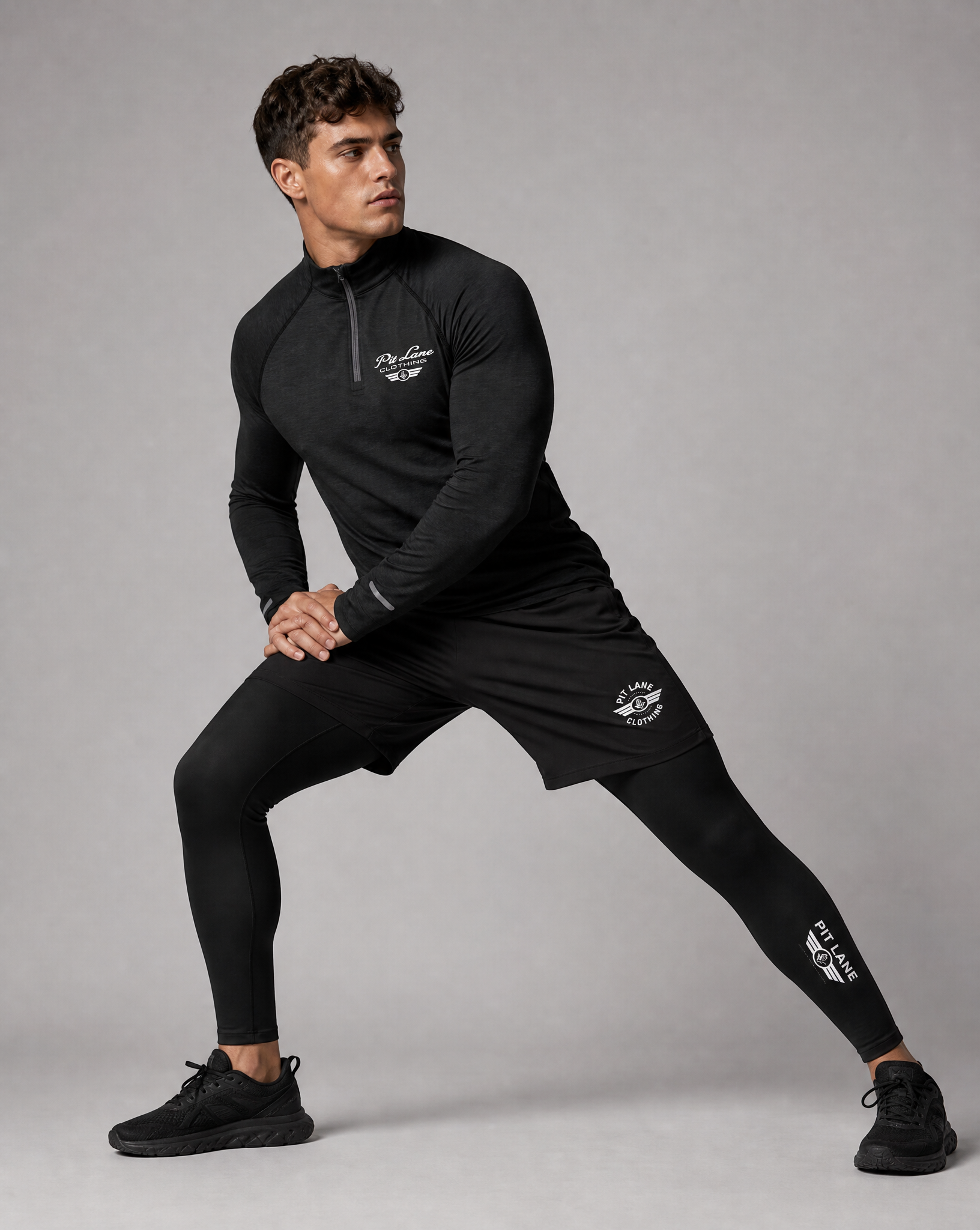 A man in a black PIT LANE CLOTHING athletic half-zip pullover, matching shorts, and full-length leggings poses in a side lunge against a solid gray studio background. The brand name 'PIT LANE CLOTHING' is printed in white on the chest, shorts, and lower leg of each garment.