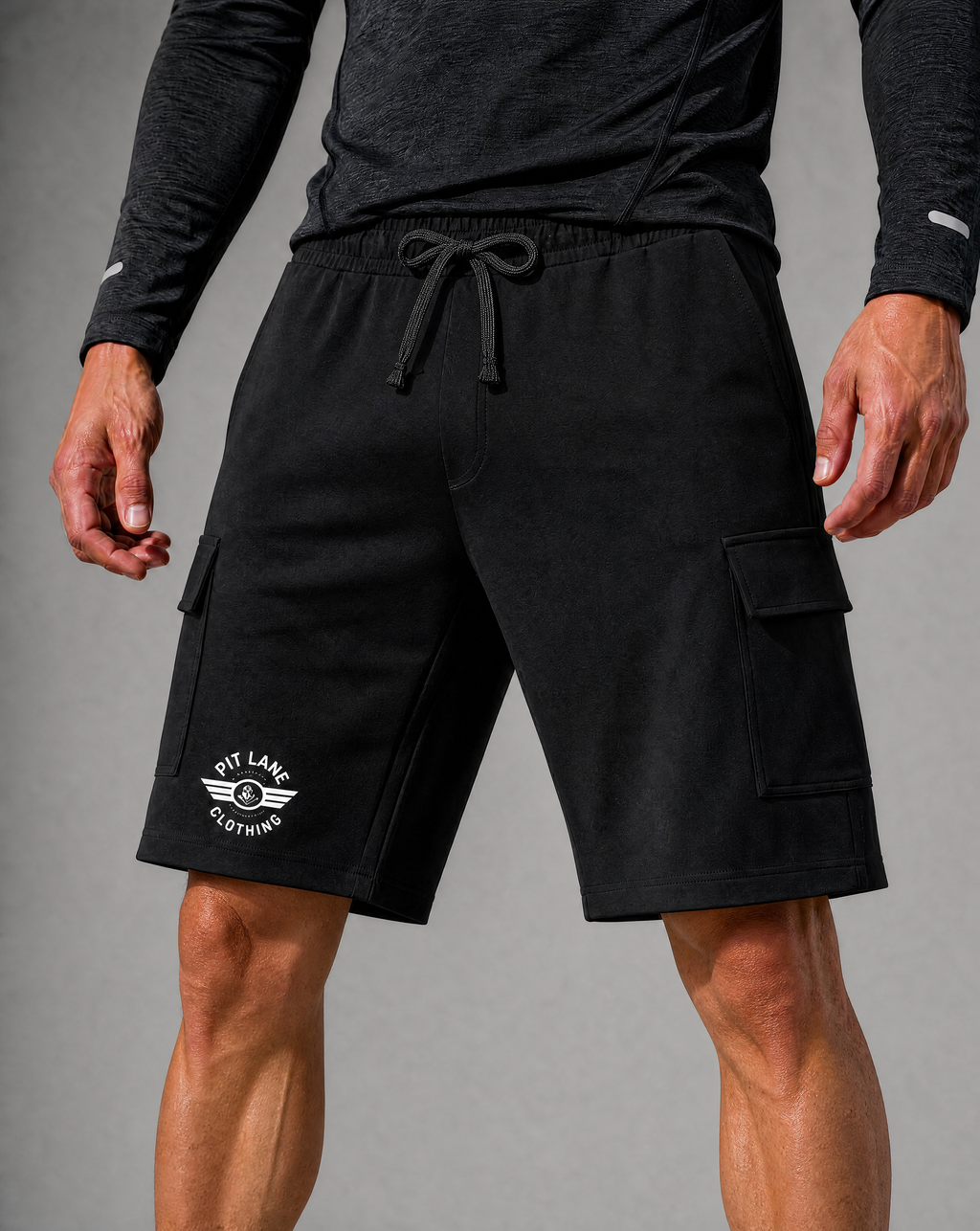 Black cargo shorts with a white PIT LANE CLOTHING printed logo on the left leg, worn by a model with a dark gray long-sleeve shirt against a gray studio background.
