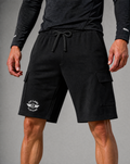 Black cargo shorts with a white PIT LANE CLOTHING printed logo on the left leg, worn by a model with a dark gray long-sleeve shirt against a gray studio background.