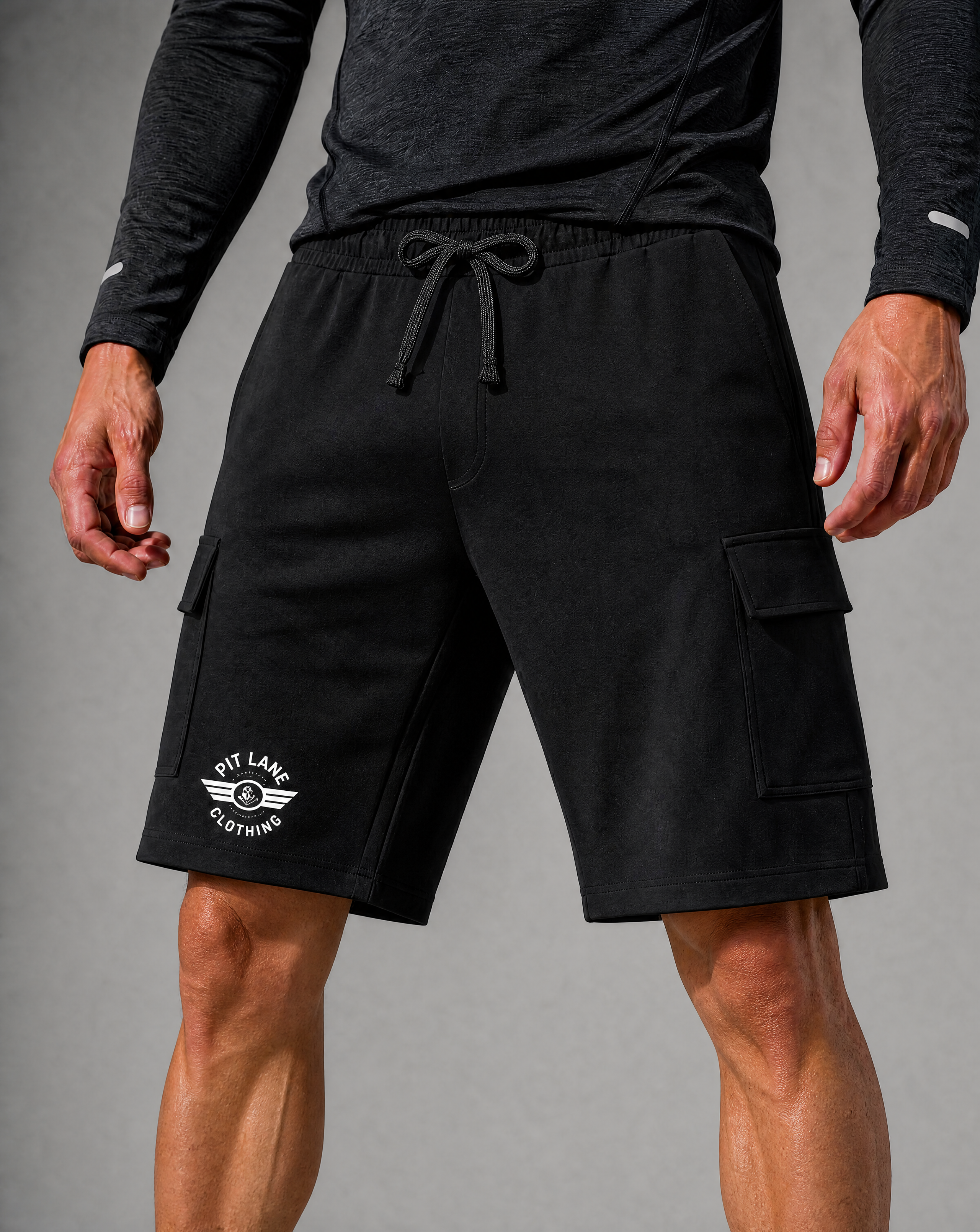 Black cargo shorts with a white PIT LANE CLOTHING printed logo on the left leg, worn by a model with a dark gray long-sleeve shirt against a gray studio background.