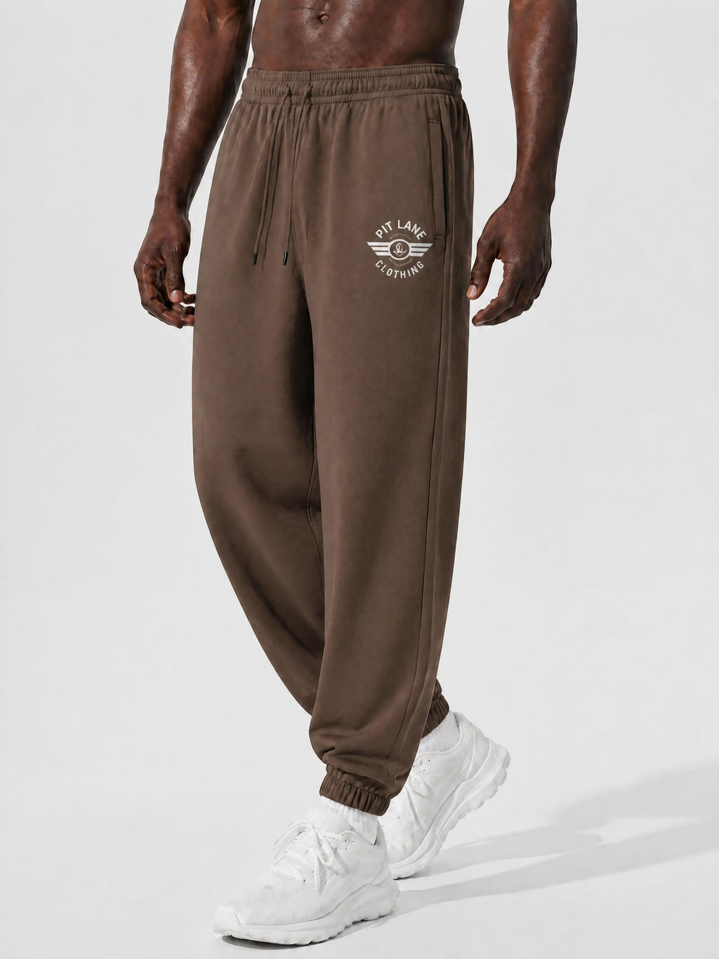 A person wearing brown jogger sweatpants with a white circular chest logo that reads 'PIT LANE CLOTHING' and white sneakers, photographed against a clean light gray studio background.