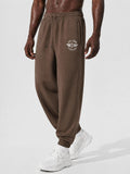 A person wearing brown jogger sweatpants with a white circular chest logo that reads 'PIT LANE CLOTHING' and white sneakers, photographed against a clean light gray studio background.