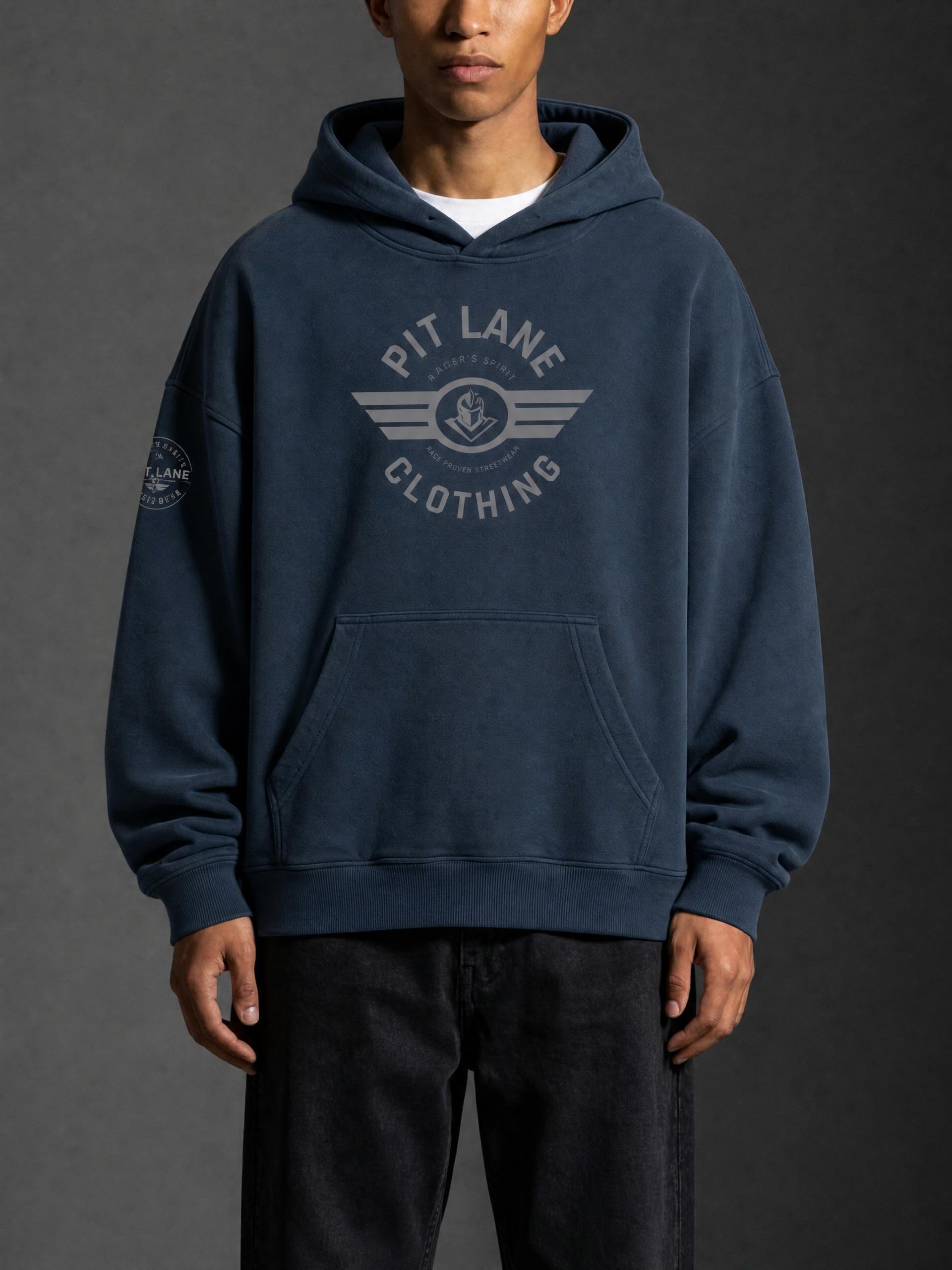 A man wearing a dark navy blue hoodie with a light gray 'PIT LANE CLOTHING' graphic on the chest, paired with black pants, against a dark gray studio background.