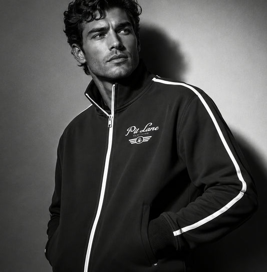 A man wearing a black zip-up track jacket with white stripe details and a white 'PIt Line Clothing' logo on the chest, photographed in a dramatic black-and-white studio setting.