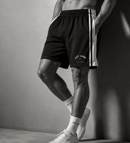 A model wearing black athletic shorts with white side stripes and a white 'PIT LAKE CLOTHING' logo on the lower left leg, styled with white crew socks and white sneakers against a gray studio background.