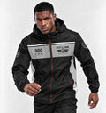 A man wearing a black and gray racing-inspired hooded jacket with '300 KPH' and 'PIT LANE' graphics, posed against a light gray studio background.