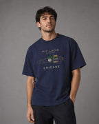 A man wearing a navy blue graphic t-shirt with a vintage race car design and 'PIT LANE CLOTHING' text, posed against a neutral gray studio background.