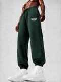 Dark green sweatpants with a white 'PIT LANE' wing logo on the left thigh, worn by a shirtless muscular model with white sneakers against a light gray studio background.