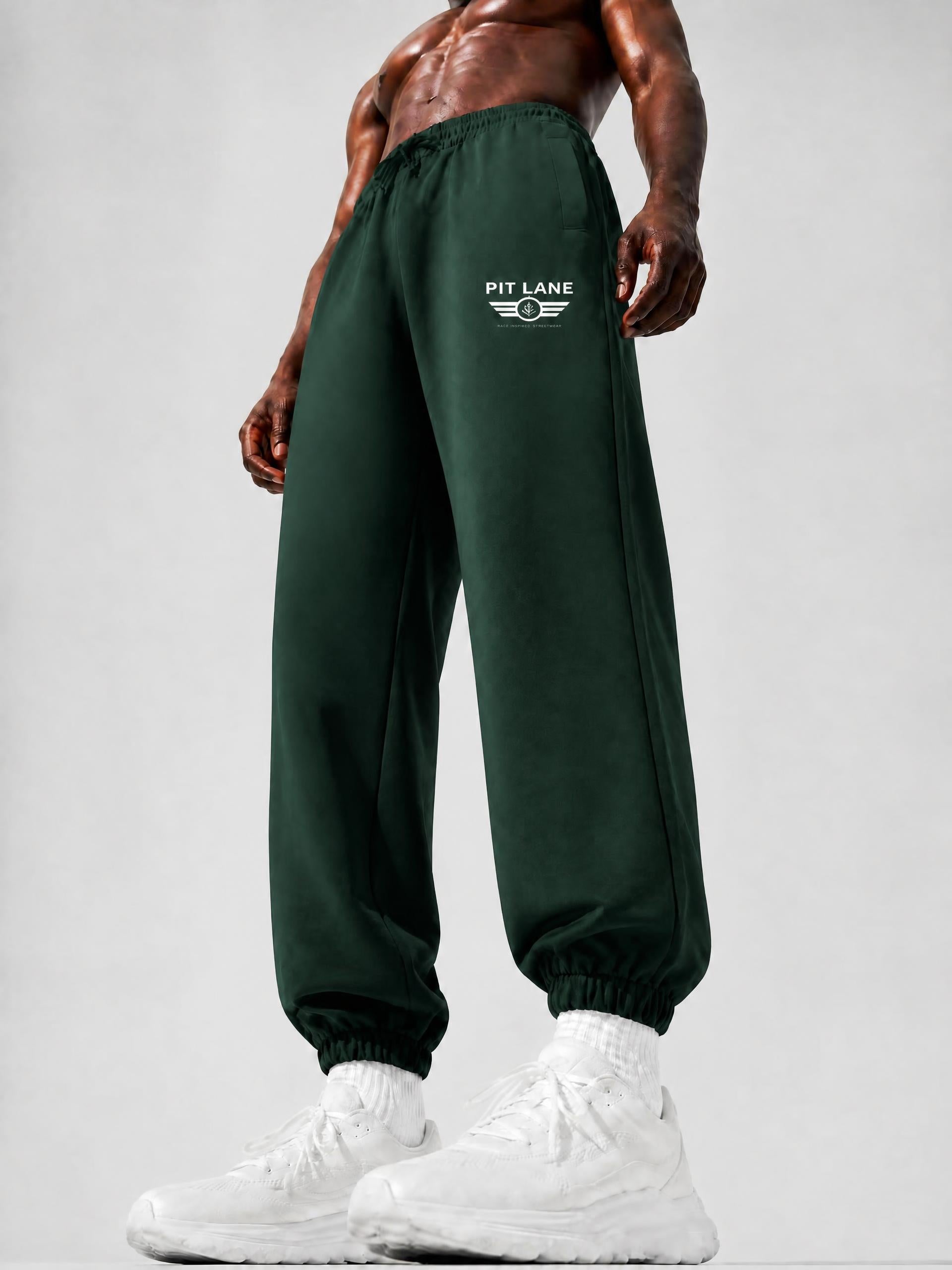Dark green sweatpants with a white 'PIT LANE' wing logo on the left thigh, worn by a shirtless muscular model with white sneakers against a light gray studio background.