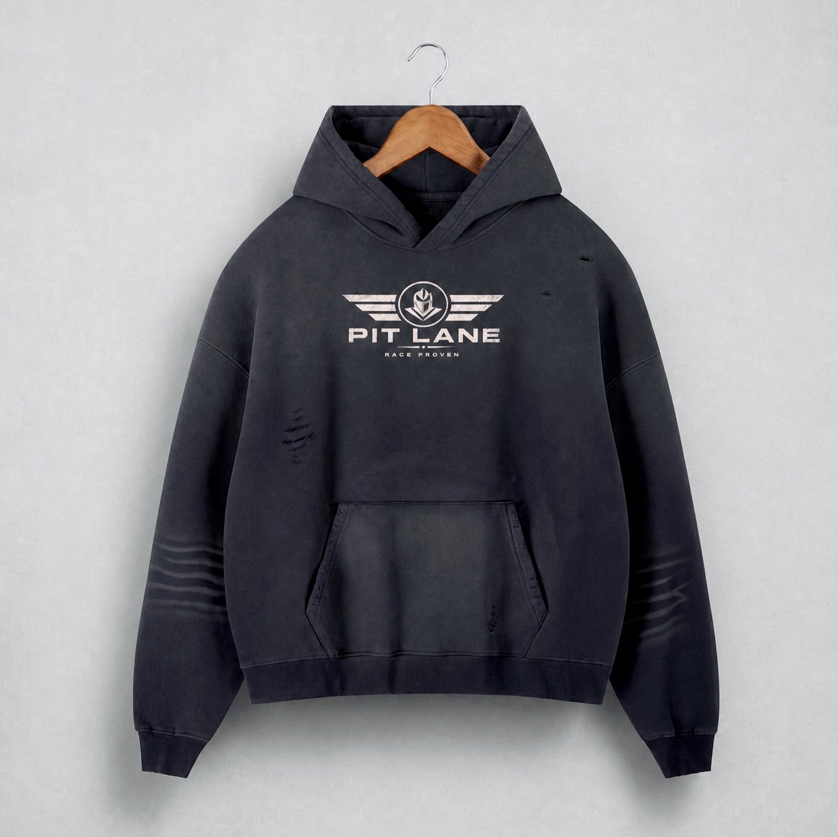 A black distressed hoodie with a white 'PIT LANE' winged logo on the chest, hanging on a wooden hanger against a light gray textured background.