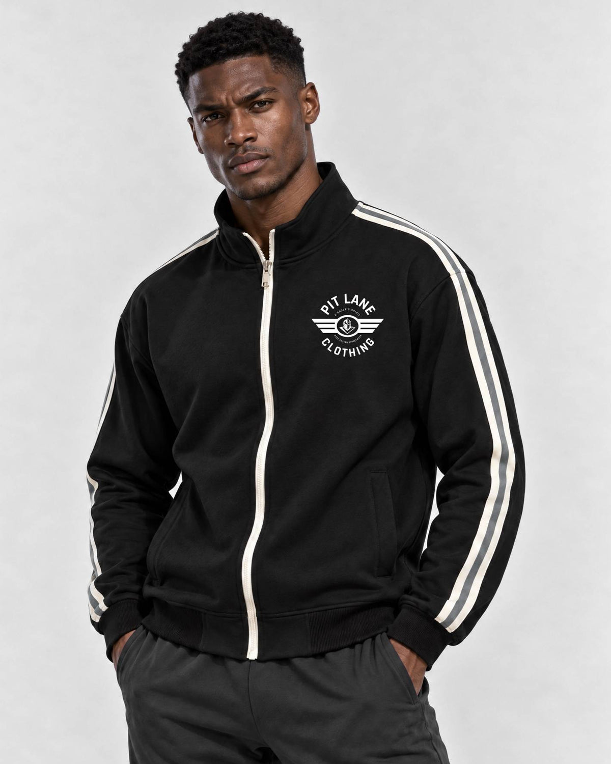 A man wearing a black zip-up track jacket with white double stripes down the sleeves and a white chest logo that reads 'PIT LANE CLOTHING', paired with dark gray sweatpants, against a light gray studio background.