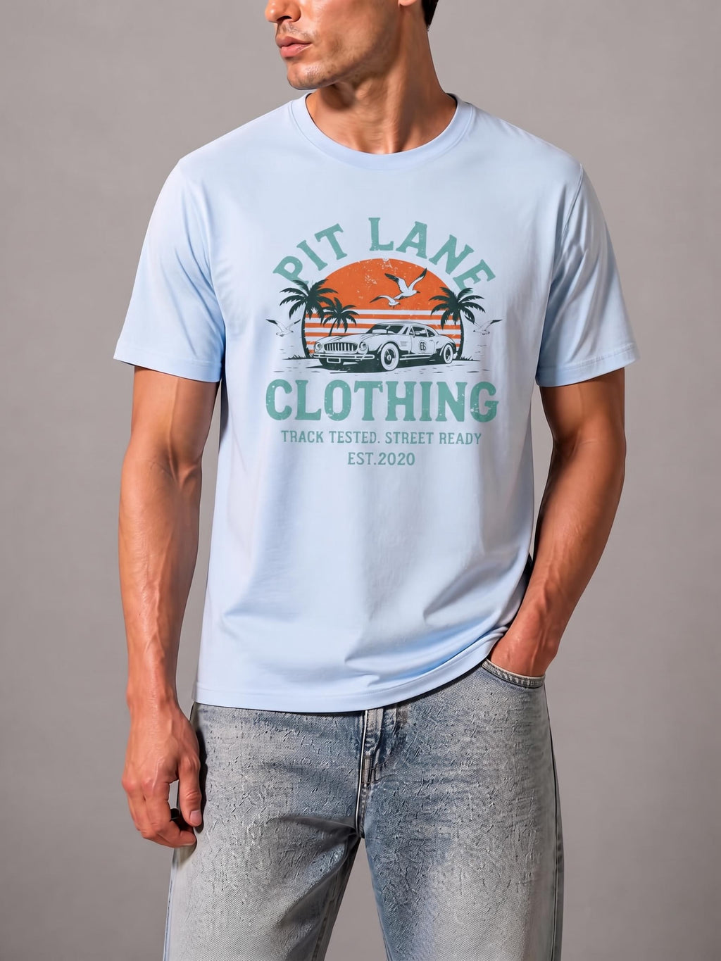 A man wearing a light blue graphic t-shirt with a retro 'Pit Lane Clothing' design, paired with light-wash jeans, against a neutral gray studio background.