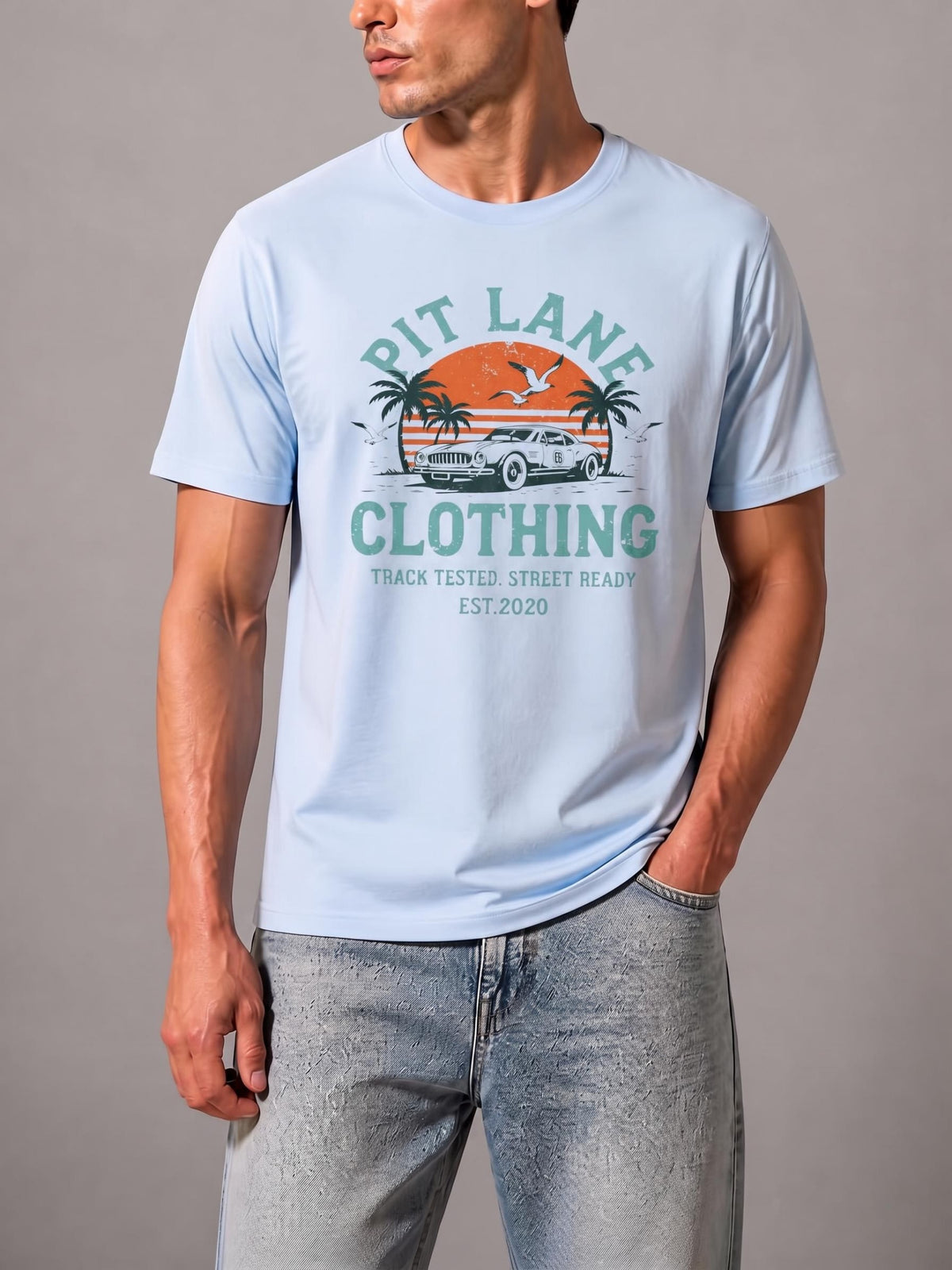 A man wearing a light blue graphic t-shirt with a retro 'Pit Lane Clothing' design, paired with light-wash jeans, against a neutral gray studio background.