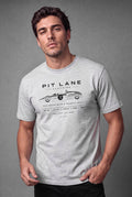 A man wearing a light gray crewneck t-shirt with a black graphic print that reads 'PIT LANE CLOTHING' and features a vintage race car illustration. The background is a solid textured gray studio backdrop.