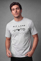 A man wearing a light gray crewneck t-shirt with a black graphic print that reads 'PIT LANE CLOTHING' and features a vintage race car illustration. The background is a solid textured gray studio backdrop.
