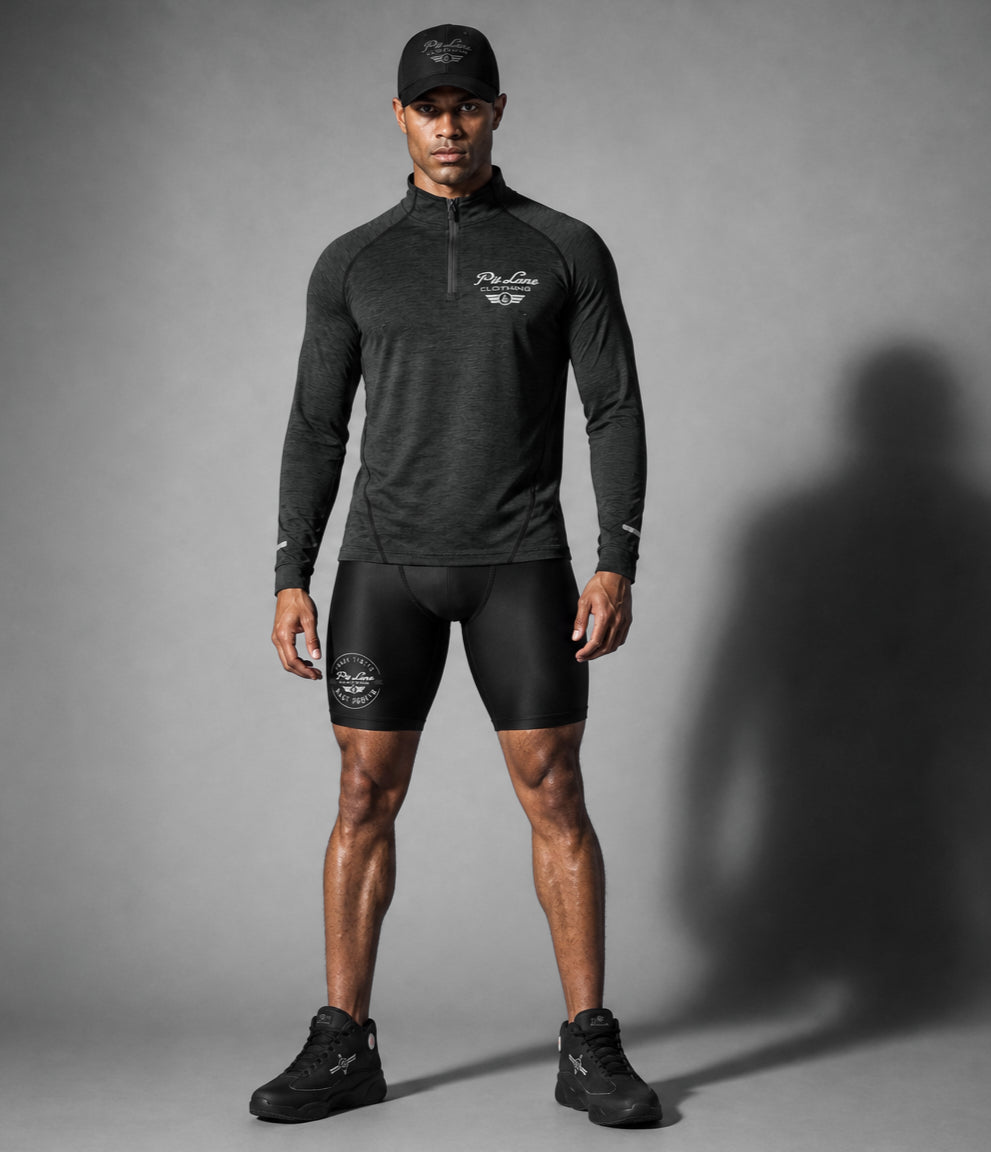 A man modeling a PIT LANE CLOTHING dark gray long-sleeve quarter-zip athletic pullover and black fitted shorts against a gray studio backdrop, with a large shadow cast to the right.