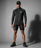 A man modeling a PIT LANE CLOTHING dark gray long-sleeve quarter-zip athletic pullover and black fitted shorts against a gray studio backdrop, with a large shadow cast to the right.