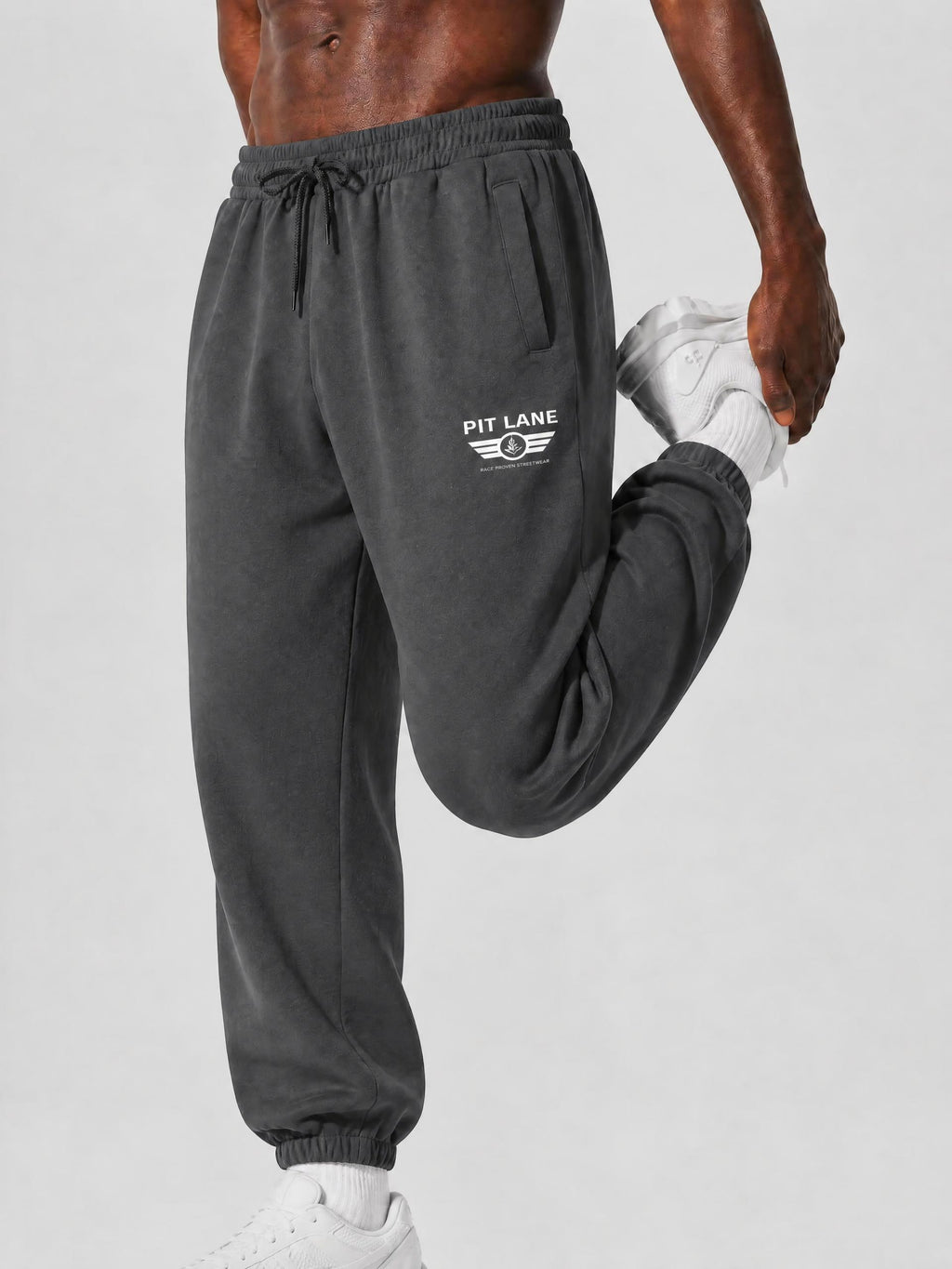 A close-up of a person wearing dark charcoal gray sweatpants with a white 'PIT LANE' wing logo on the left thigh, stretching their leg upwards while holding a white sneaker against a light gray studio background.