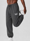 A close-up of a person wearing dark charcoal gray sweatpants with a white 'PIT LANE' wing logo on the left thigh, stretching their leg upwards while holding a white sneaker against a light gray studio background.