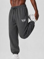 A close-up of a person wearing dark charcoal gray sweatpants with a white 'PIT LANE' wing logo on the left thigh, stretching their leg upwards while holding a white sneaker against a light gray studio background.