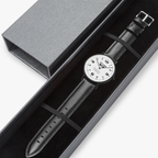 A Pit Lane wristwatch with a black leather strap, black case, and a white dial with black Arabic numerals, shown against a light gray background.