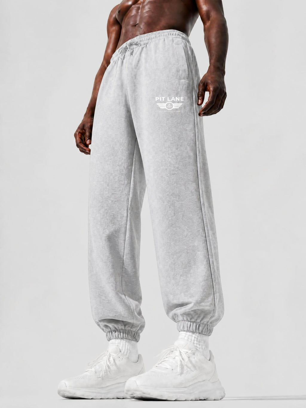 A model wearing light gray heather jogger sweatpants with a white 'PIT LANE' logo on the left thigh, paired with white sneakers, shown from the waist down against a light gray studio background.