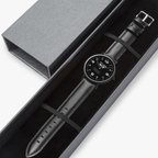 A luxury PIT LANE CLOTHING watch with a black leather strap and a black dial, presented inside a dark gray gift box with its lid placed beside it on a white background.