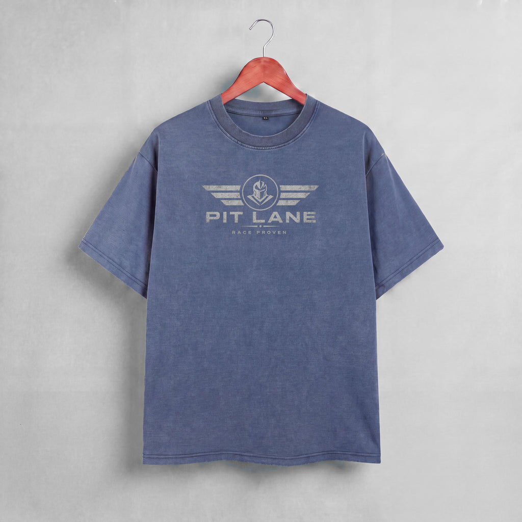 A washed slate blue short-sleeve t-shirt hanging on a wooden hanger against a textured gray wall. The shirt features a distressed white graphic on the chest with the text 'PIT LANE' and 'RACE PROVEN' beneath it.