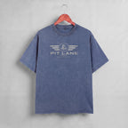 A washed slate blue short-sleeve t-shirt hanging on a wooden hanger against a textured gray wall. The shirt features a distressed white graphic on the chest with the text 'PIT LANE' and 'RACE PROVEN' beneath it.
