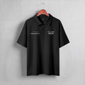 A dark navy blue short-sleeve polo shirt hanging on a wooden hanger against a light gray textured wall. The shirt features white embroidered graphics on the chest, including 'PIT LANE' with wings on the left chest and a script logo on the right chest.