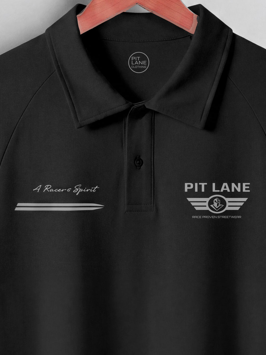 A black polo shirt hanging on a wooden hanger, featuring 'PIT LANE' graphics on the chest with a winged logo and 'A Racing Spirit' script on the opposite side.
