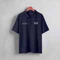 A dark navy blue short-sleeve polo shirt hanging on a wooden hanger against a light gray textured wall. The shirt features white embroidered graphics on the chest, including 'PIT LANE' with wings on the left chest and a script logo on the right chest.