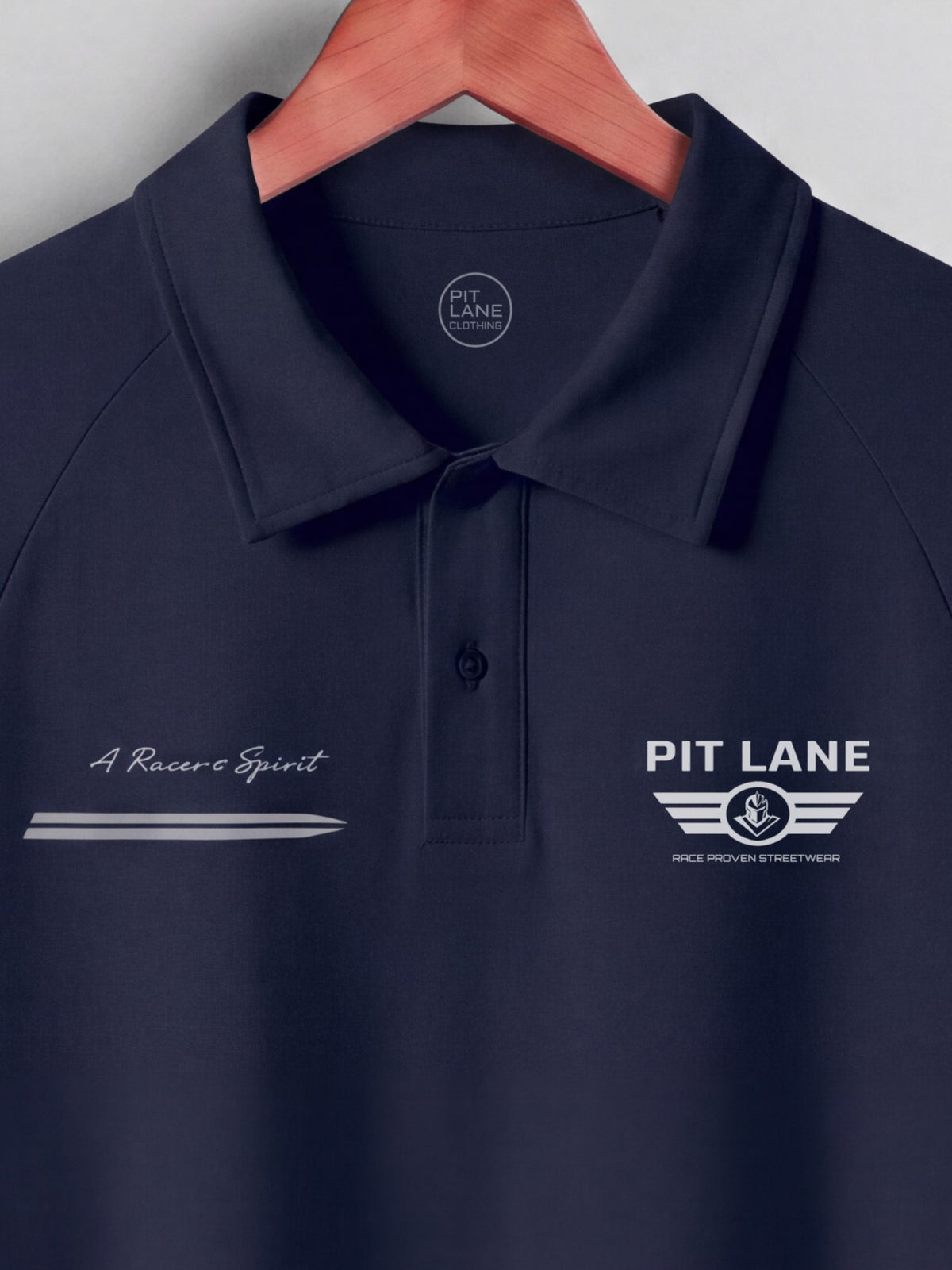 A navy blue polo shirt on a wooden hanger, featuring white embroidered logos and text on the chest. The left chest reads 'A Racer's Spirit' with a stylized arrow, and the right chest reads 'PIT LANE' with winged stripes and 'SPICE PREMIUM STREETWEAR'.