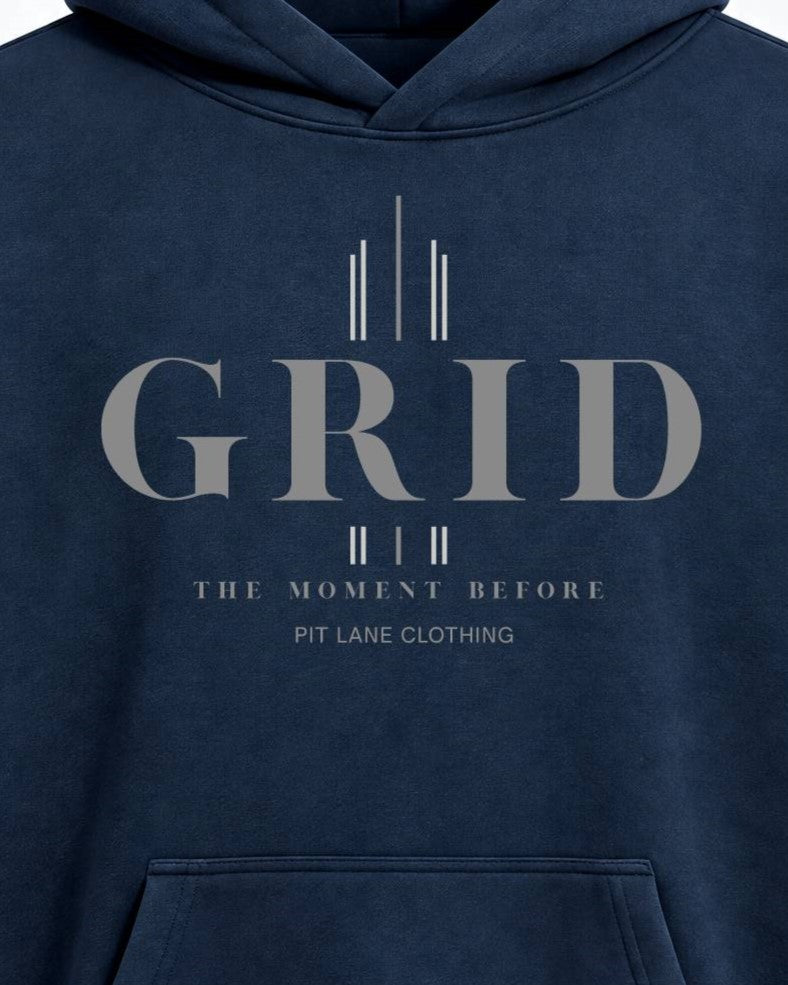 Close-up of a navy blue hoodie featuring a light gray graphic print on the chest with the word 'GRID' and smaller text reading 'THE MOMENT BEFORE' and 'PIT LANE CLOTHING'.