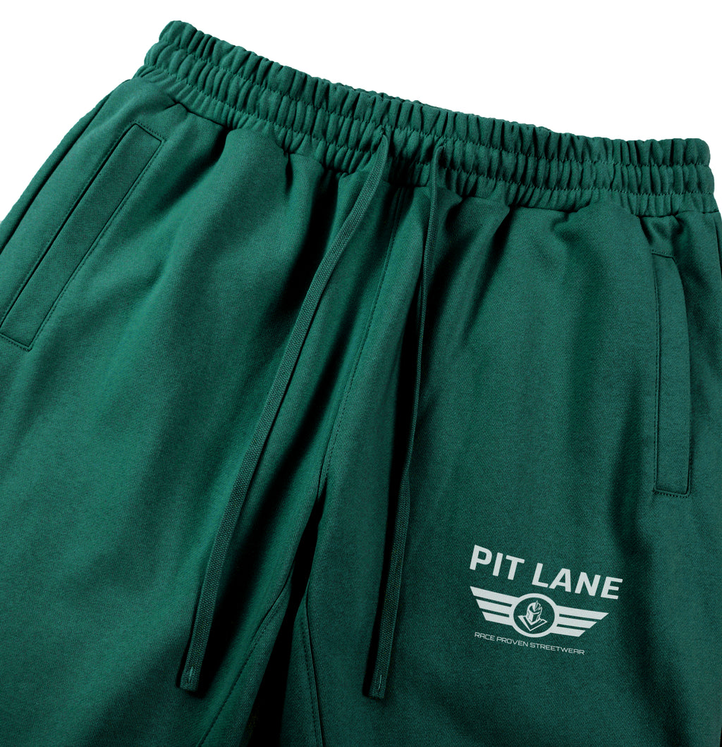 Close-up of dark green athletic shorts with an elastic waistband, drawstrings, side pockets, and a white 'PIT LANE' logo printed on the lower left leg.