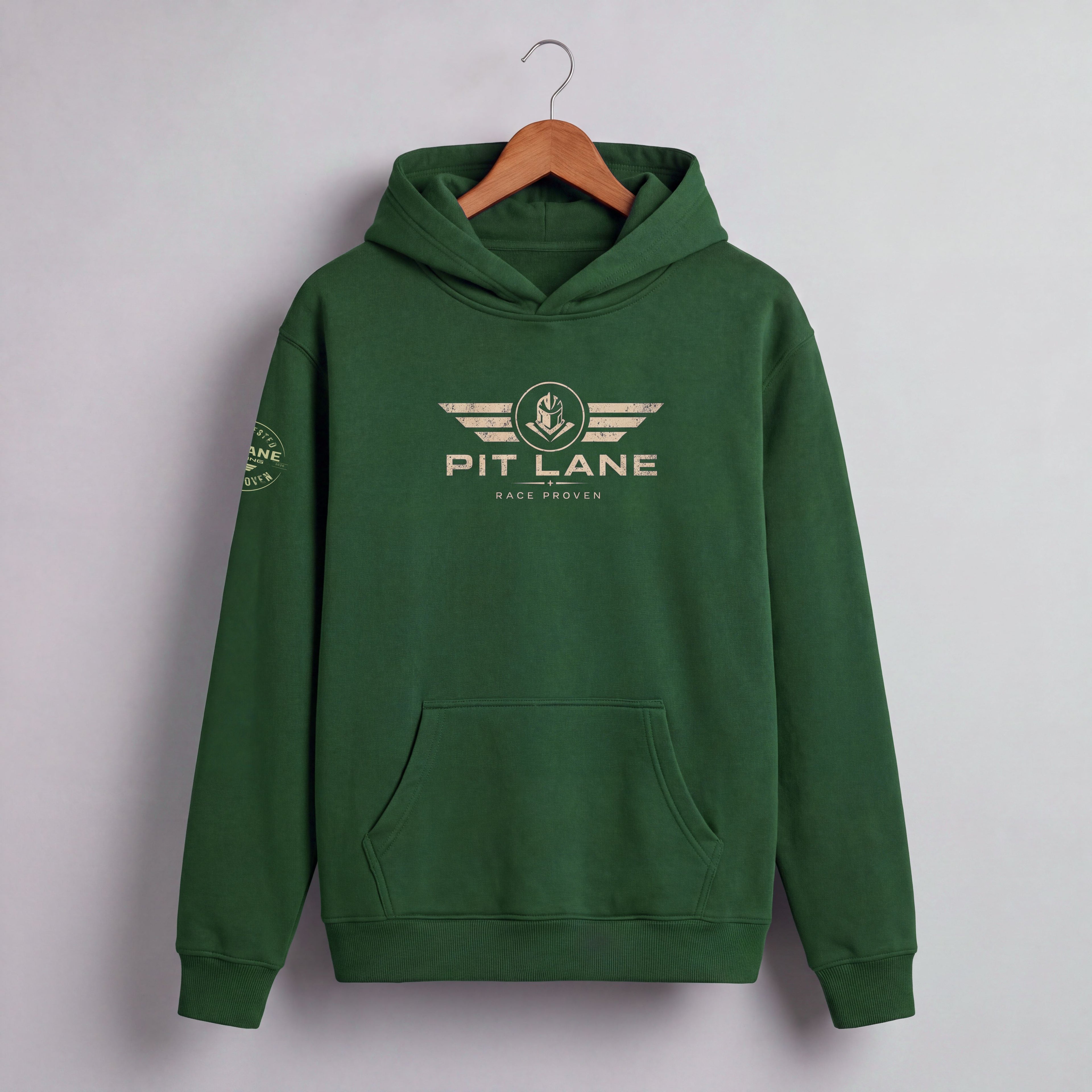 A dark green pullover hoodie hanging on a wooden hanger against a light gray wall. The hoodie features a beige graphic on the chest with the text 'PIT LANE' and 'RACE PROVEN' beneath it, and a matching graphic on the left sleeve.