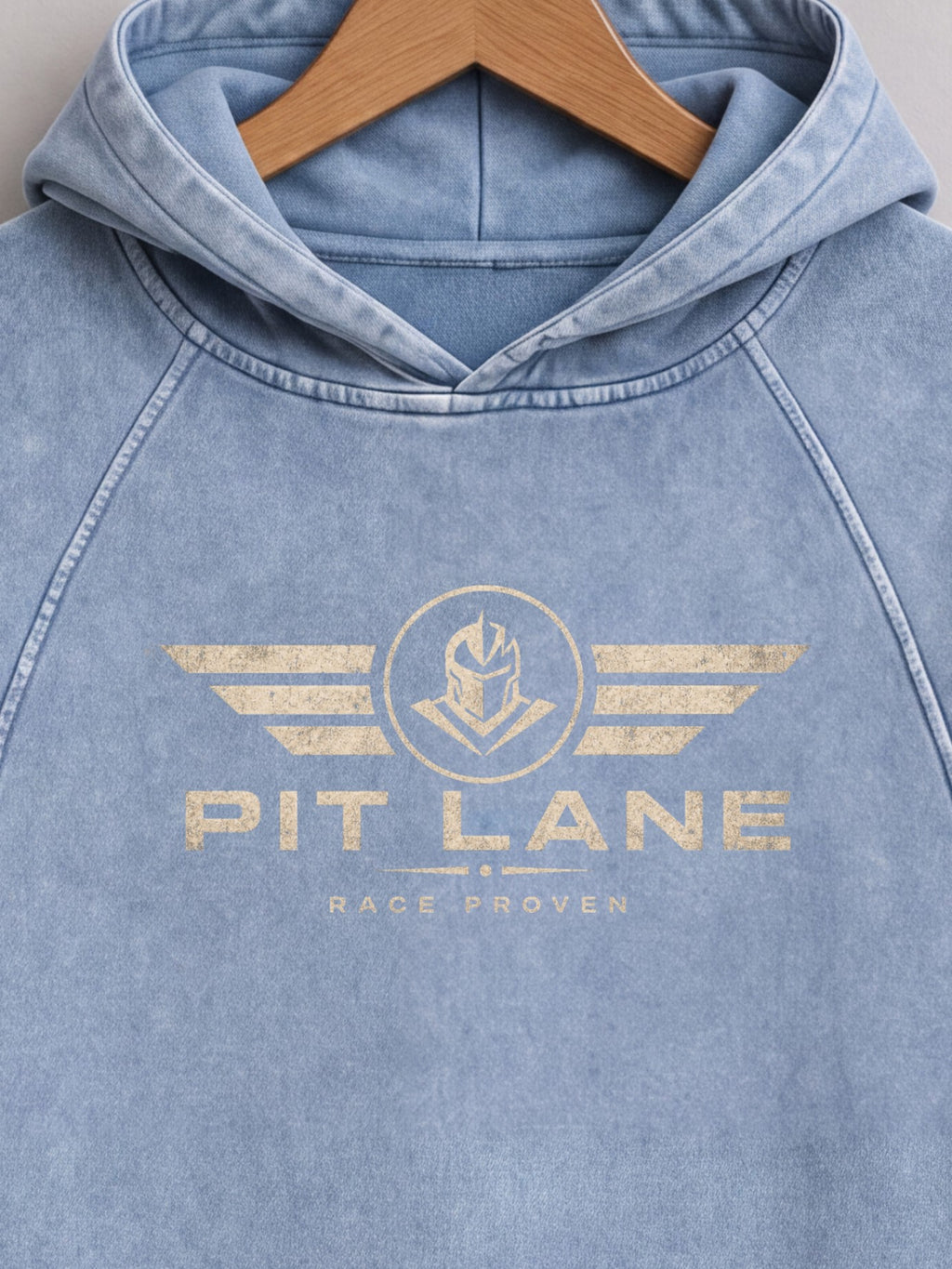 A light blue washed hooded sweatshirt hanging on a wooden hanger, featuring a distressed gold graphic on the chest with wings, a helmet emblem, and the text 'PIT LANE' and 'RACE PROVEN'.