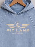 A light blue washed hooded sweatshirt hanging on a wooden hanger, featuring a distressed gold graphic on the chest with wings, a helmet emblem, and the text 'PIT LANE' and 'RACE PROVEN'.