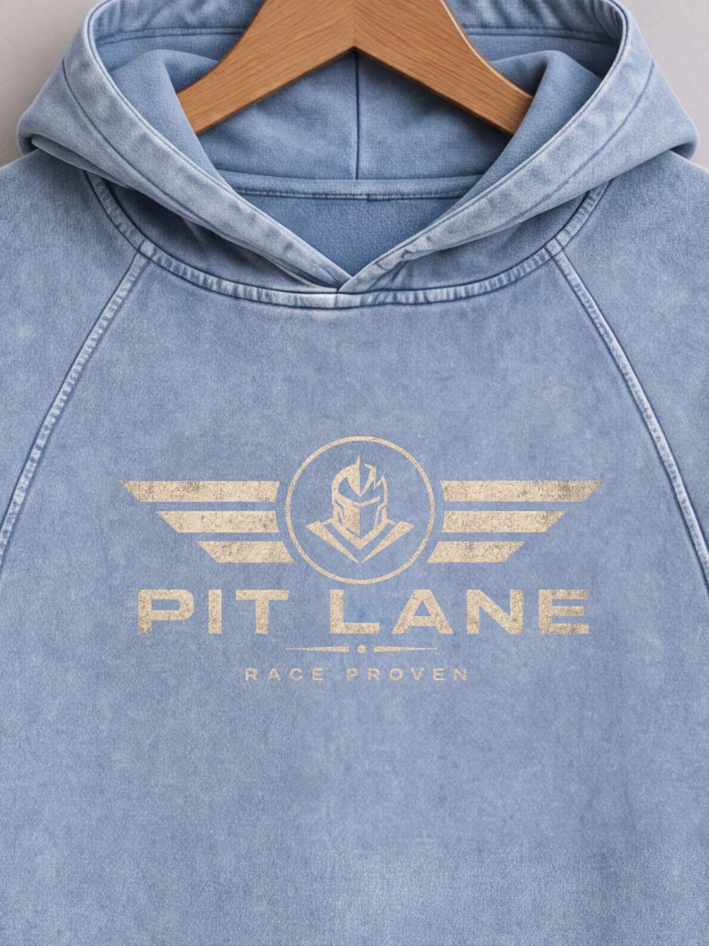 A light blue washed hooded sweatshirt hanging on a wooden hanger, featuring a distressed gold graphic on the chest with wings, a helmet emblem, and the text 'PIT LANE' and 'RACE PROVEN'.