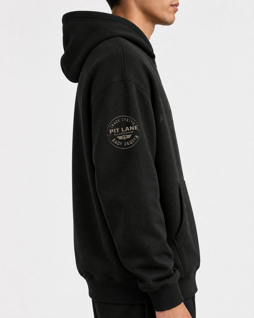 A man wearing a black pullover hoodie with a circular 'PIT LANE' logo printed on the left sleeve, shown in a side profile against a light gray background.