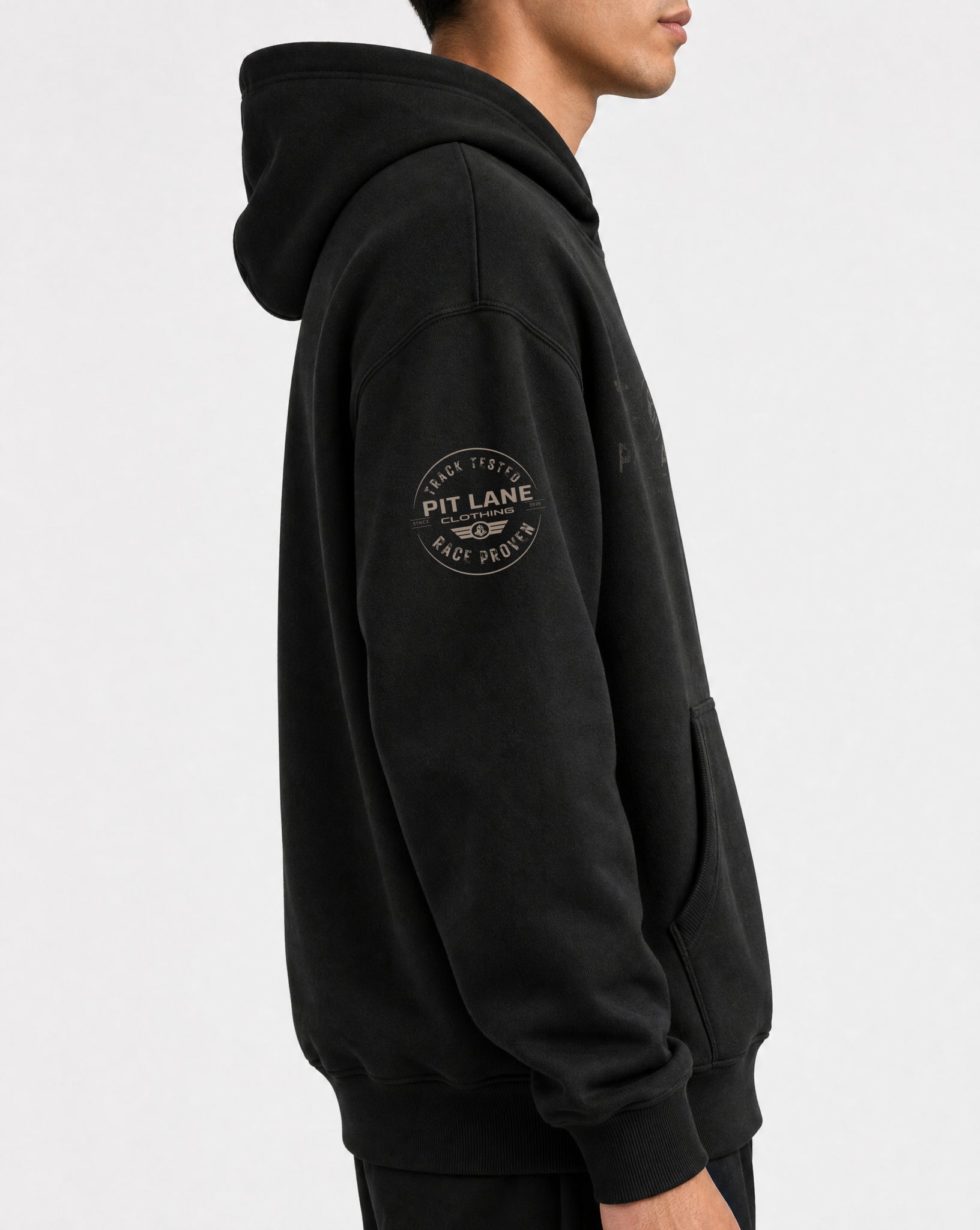 A man wearing a black pullover hoodie with a circular 'PIT LANE' logo printed on the left sleeve, shown in a side profile against a light gray background.