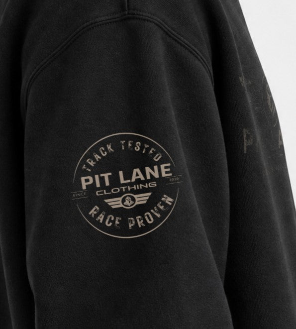 Close-up of a black sweatshirt sleeve featuring a circular distressed graphic on the upper arm that reads 'PIT LANE CLOTHING' with 'TRACK TESTED' and 'RACE PROVEN'.